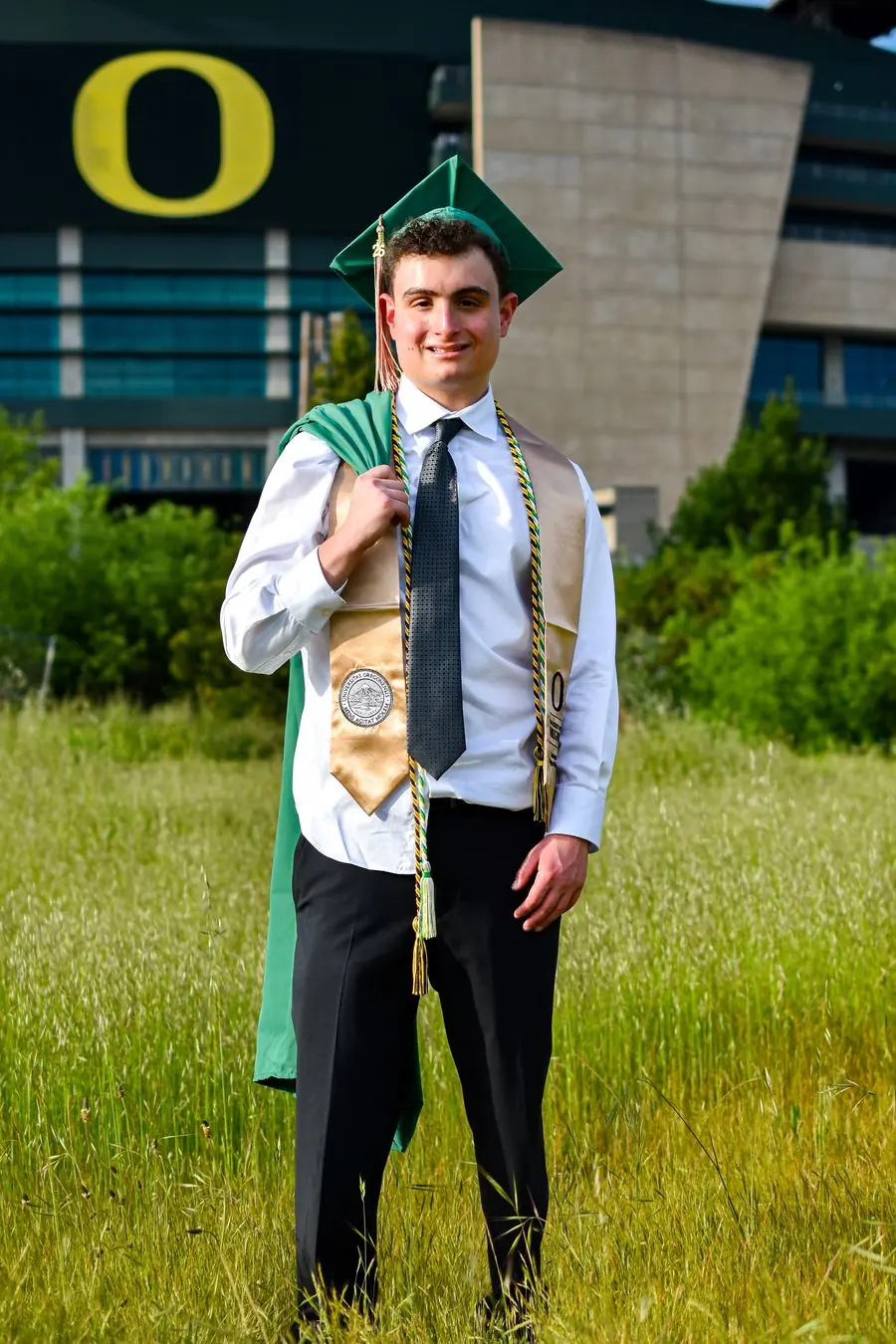 Graduate holding his cap and gown over his shoulder in front of Autzen Stadium