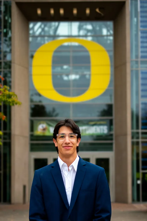 Headshot of a smiling man in clear glasses and a navy blazer beneath the University of Oregon O