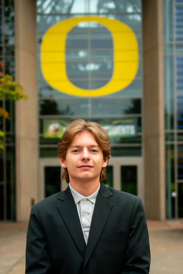 Headshot of a man in a black blazer and gray polo beneath the University of Oregon O