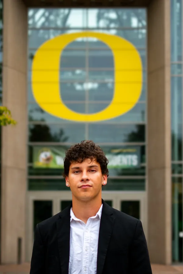 Headshot of a curly-haired man in a black blazer beneath the University of Oregon O