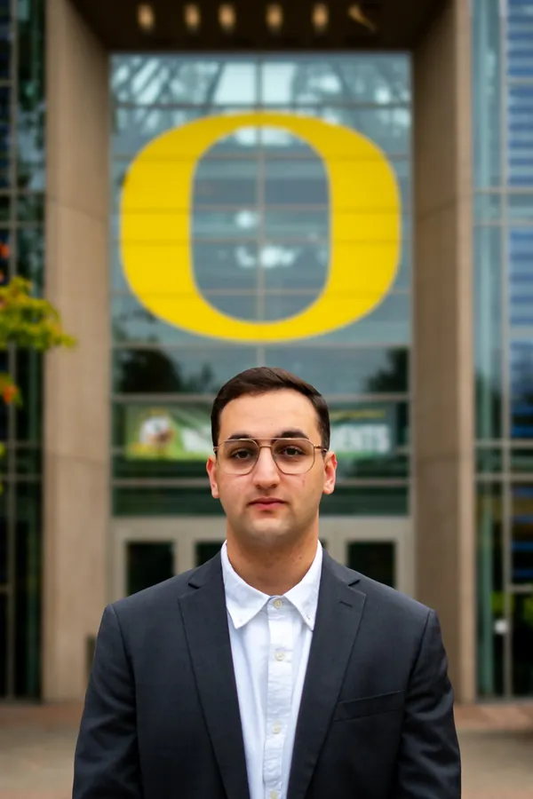 Headshot of a man in gold-rimmed glasses and a charcoal suit beneath the University of Oregon O